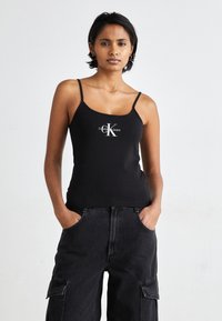 Black tank top with spaghetti straps featuring a white CK logo. Worn with high-waisted, loose-fitting black cargo pants. Smooth fabric.