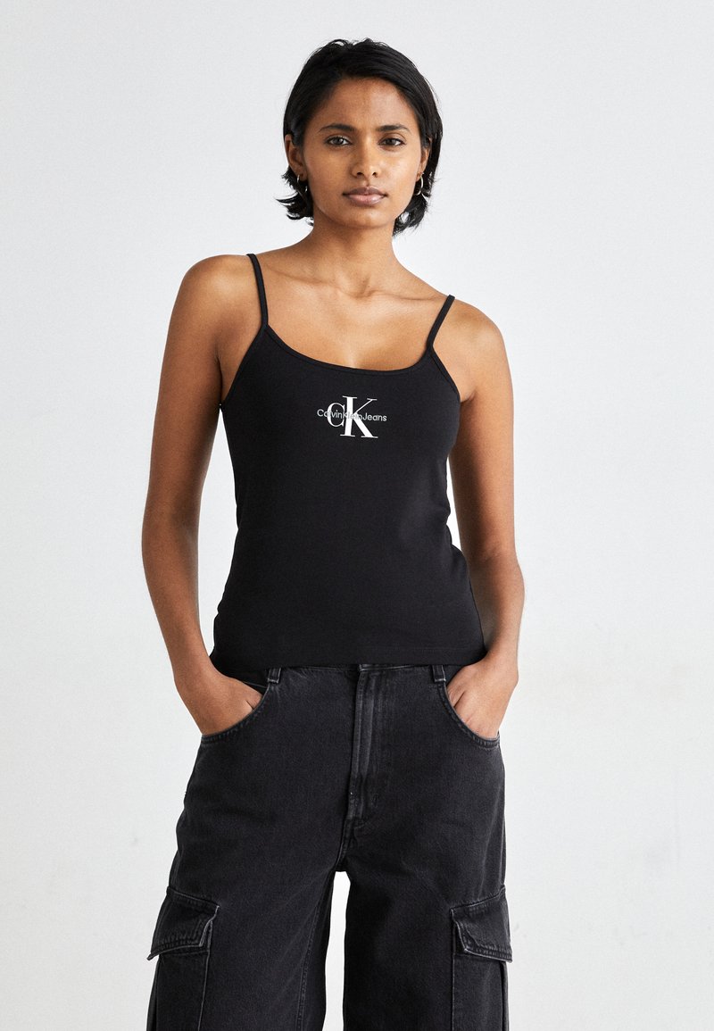 Black tank top with spaghetti straps featuring a white CK logo. Worn with high-waisted, loose-fitting black cargo pants. Smooth fabric.