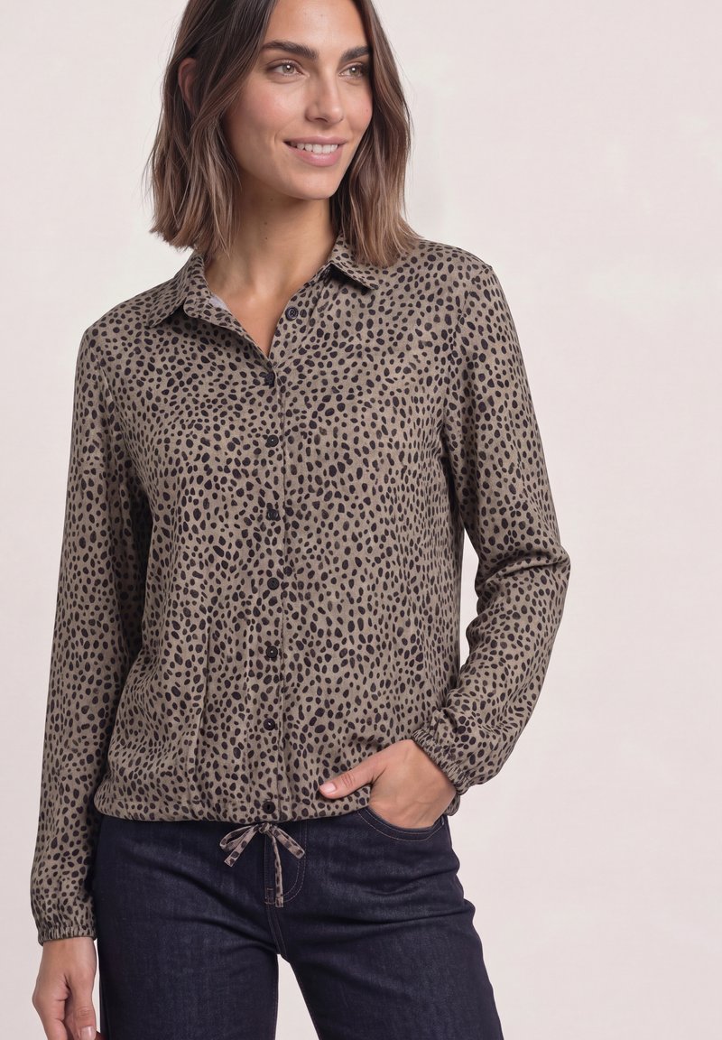 Femme portant une chemise à manches longues boutonnée avec un motif tacheté marron et noir et un jean foncé, main dans la poche, souriant légèrement.
