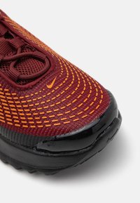 Nike trkačka cipela s gornjim dijelom od mrežastog materijala u tamnocrvenoj i narančastoj boji, teksturiranom površinom, crnim gumenim potplatom i zaobljenim vrhom s akcentima od šivanja.
