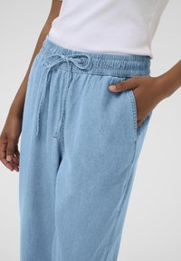 Lyseblå denimbukser med elastisk talje og snor, med sidelommer og afslappet pasform, vist fra taljen og ned.
