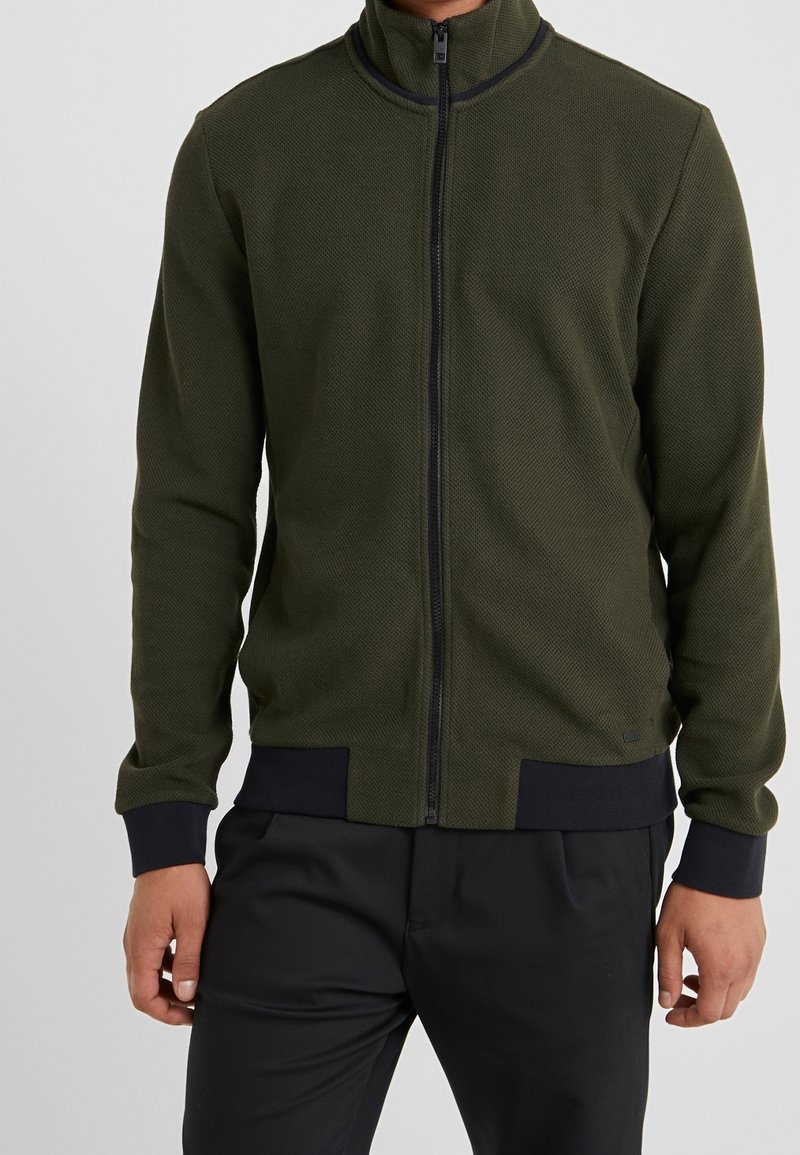 Grüne Reißverschlussjacke mit erhabener Textur, hohem Kragen sowie schwarzen, gerippten Bündchen und Saum. Verfügt über einen vorderen Reißverschluss und dezente Markenkennzeichnung.