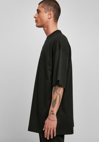 Zwarte oversized t-shirt met korte mouwen, gemaakt van katoen. Heeft een ronde hals. Model heeft tatoeages en draagt een zilveren armband om de pols.