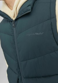 Padded vest in dark teal with a high collar, featuring a front zipper and embossed "Originals Studio" logo. Smooth fabric texture.