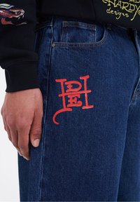 Mörkblå denimjeans har en röd broderad logotyp på vänster lår, med en rak ben-design och traditionell femfickstil.