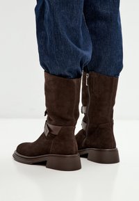 Botas de tobillo de ante marrón con tacón ancho, cremallera lateral y detalles decorativos de tiras de cuero. Acabado texturizado, punta redondeada.