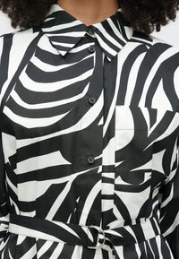 Juodai baltas mygtukais užsegamas marškinėlis su zebro dryžiais, apykakle, krūtinės kišene ir derančia diržu su sidabrine D formos sagtimi.