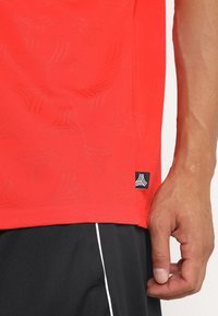 Oikea käsi vartalon vieressä, ja yllä kirkkaan punainen kuviollinen paita, jossa on pieni musta logotunniste, sekä mustat housut, joissa on valkoinen raita.