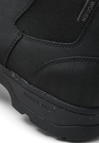Bota negra con parte superior de tela texturizada, costuras reforzadas y suela de goma con un patrón de relieve. Logo "Natural Soft" visible en el lateral.