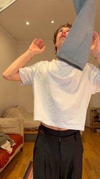 Άτομο που φοράει λευκό crop top και μαύρο παντελόνι σηκώνει ένα γκρι πουλόβερ πάνω από το κεφάλι μέσα σε ένα δωμάτιο με ξύλινο πάτωμα και έπιπλα.