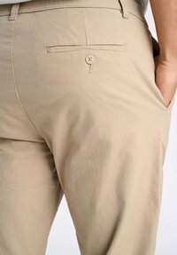 Pantalons beige clair avec une texture lisse, dotés d'une poche arrière et d'une fermeture à un seul bouton. Design épuré, détails de couture subtils.