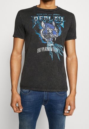 Homme portant un t-shirt noir à motif d'un tigre bleu rugissant avec des éclairs bleus, associé à un jean bleu.