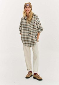 Blondynka w brązowej czapce, oversize'owej koszuli w kratę, jasnobeżowych spodniach, bordowych skarpetkach i beżowych butach trekkingowych, stojąca z rękami w kieszeniach.