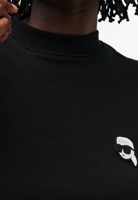 Czarna bluza z okrągłym dekoltem i gładką teksturą, z kolorową naszywką logo postaci w okularach przeciwsłonecznych na piersi.