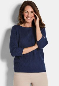 Dunkelblau gestrickter Pullover mit Dreiviertelärmeln, der ein strukturiertes Muster sowie gerippte Bündchen und Saum aufweist. Getragen mit hellbeigen Hosen.