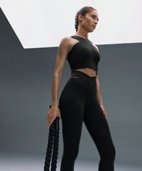 Top sportiv negru cu design asimetric, asociat cu colanți negri cu talie înaltă. Modelul ține o frânghie de antrenament neagră.