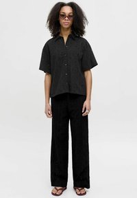 Mujer con cabello rizado vistiendo una camisa negra de botón con ojales y pantalones anchos a juego, sandalias moradas y gafas de sol, de pie frente a un fondo blanco.