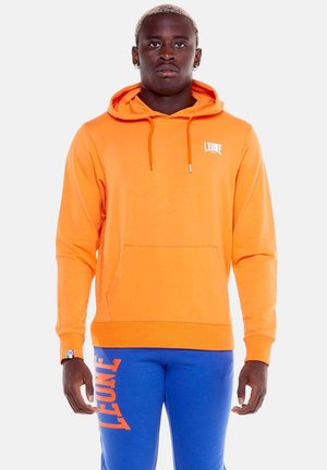 Mann, der nach vorne schaut und einen orangen Hoodie mit weißem Logo sowie blaue Jogginghose mit großem orangem Text am linken Bein trägt.