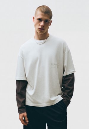 Jeune homme aux cheveux courts portant une chemise blanche à manches courtes par-dessus une chemise noire à manches longues, un pantalon noir et un collier à chaîne fine.