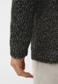 Maglione chunky a maglia grigia scura con sfumature più chiare, maniche lunghe, polsini a costine e vestibilità rilassata, mostrato con una mano appoggiata vicino all'orlo.