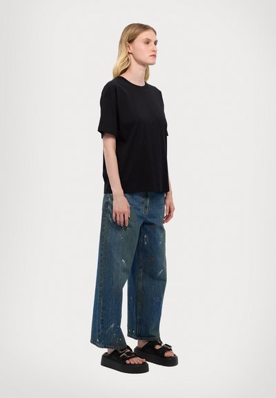 T-shirt noir à manches courtes associé à un jean en denim délavé, ample, avec des éclaboussures de peinture ; le modèle porte des sandales noires massives.