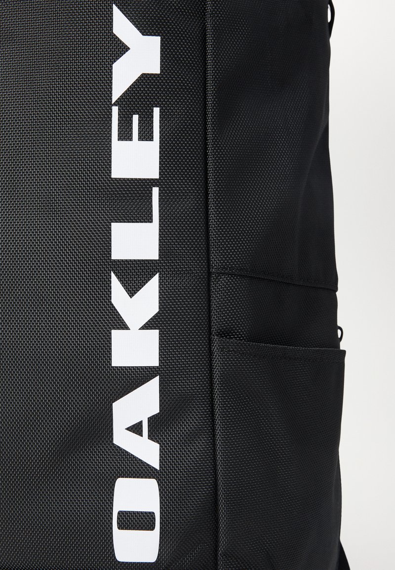 Zwarte rugzak met een gestructureerde stof. Beschikt over een opvallend wit "OAKLEY" logo, een zijvak en verstelbare banden voor functionaliteit.