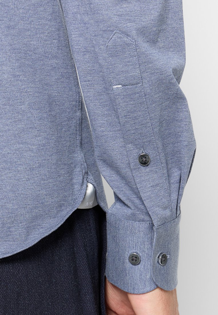 Camisa de manga larga azul con tejido texturizado, que presenta cierres de botones negros en las mangas y un pequeño bolsillo en el pecho.