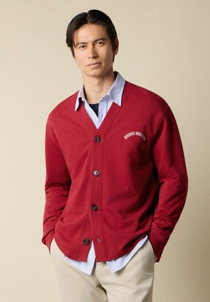 Uomo che indossa un cardigan rosso con bottoni sopra una camicia a righe blu e pantaloni beige, in piedi con le mani nelle tasche contro uno sfondo neutro.
