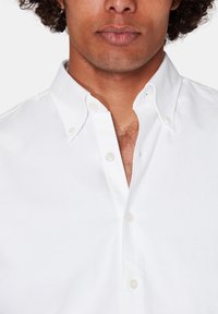 Camicia bianca a maniche lunghe in cotone, con colletto tradizionale, dettagli dei bottoni in vista e una texture liscia.