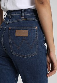 Osoba v modrých džínách Wrangler se střiženým W na zadní kapse a koženou nášivkou v barvě tan, bílá košile zasunutá dovnitř, ruka uvolněná podél těla.