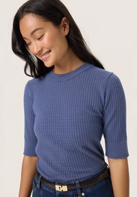 Ribbed knit short-sleeve top in soft blue. Features a round neckline and textured pattern. Paired with denim and a brown belt.