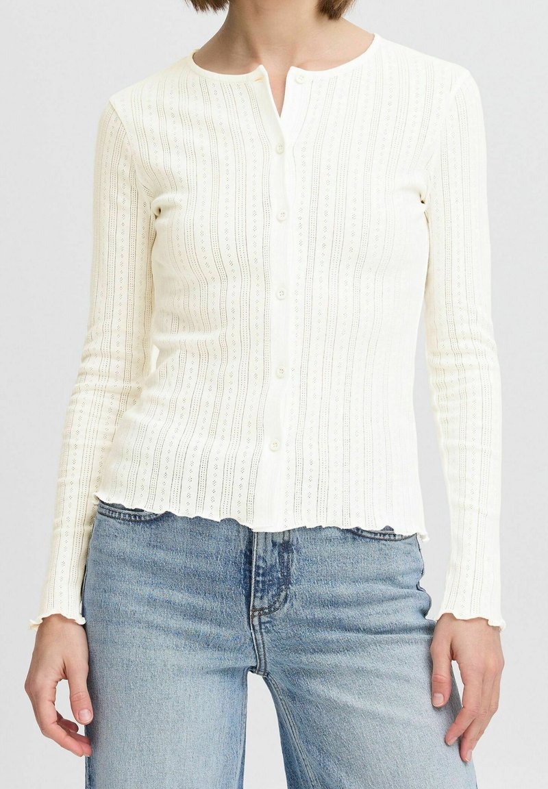 Biała kardigan z długim rękawem, w pionowy wzór, okrągłym dekoltem, z zapięciem na guziki i falbaniastym dołem. Tkanina ma prążkowaną strukturę.