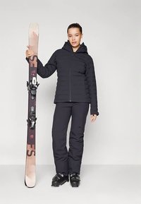 Γυναίκα με μαύρο σκι σάκο και παντελόνι, κρατώντας ένα σκι σε όρθια θέση, φορώντας μαύρες μπότες σκι, που στέκεται σε ουδέτερο γκρίζο φόντο.