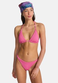 Bikini rosa a coste con top triangolare e spalline sottili, abbinato a slip a vita alta coordinati. La modella indossa un foulard viola con fantasia.