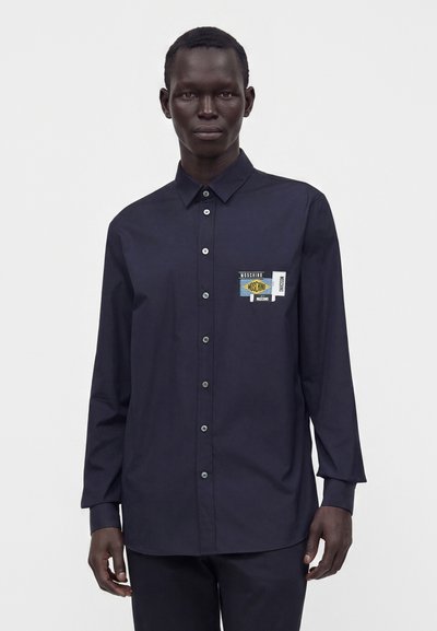 Homme portant une chemise bleu marine foncé boutonnée avec un patch logo Moschino coloré sur la poitrine gauche sur fond blanc uni.
