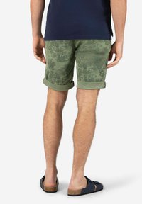 Man wearing rolled-up green patterned shorts, navy blue shirt, and black open-back sandals, standing against a plain background.