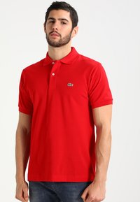 Červená polo tričko z bavlny s klasickým golierom, krátkymi rukávmi a malým zeleným logom krokodíla na ľavej hrudi.