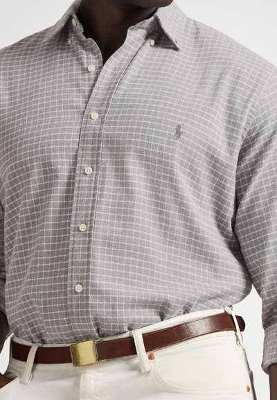 Sivo karo srajco z gumbi iz tkanine, na prsih je majhen logotip, oblečena s rjavim usnjenim pasom z zlatim zaponko.