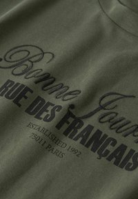 Olijfgroen stof met zwarte tekst die "Bonne Faur," "RUE DES FRANÇAIS," en "ESTABLISHED 1992 75011 PARIS" zegt.