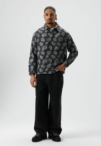 Zwart overhemd met patronen en witte, spiraalvormige ontwerpen, met kraag en lange mouwen, gecombineerd met losse zwarte jeans en zwarte plateauzolen.