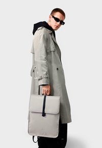 Grå veske med klaffelukking, mørk stropp og læraktig tekstur, holdt av en person i en lys trenchcoat og svart hettegenser.