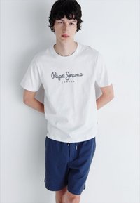 Balta medvilninė marškinėliai su pilka "Pepe Jeans London" logotipu, derinami su tamsiai mėlynomis šortais. Apranga pasižymi laisvu kirpimu ir paprastu dizainu.