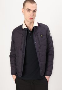 Veste matelassée marine avec un col blanc contrasté, fermeture éclair à l'avant et deux poches zippées. Présente un écusson logo sur la manche gauche.