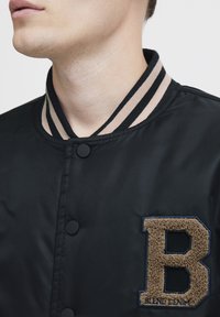 Black satin bomber jacket with a beige striped ribbed collar. Features a textured brown fabric letter "B" patch and snap buttons.
