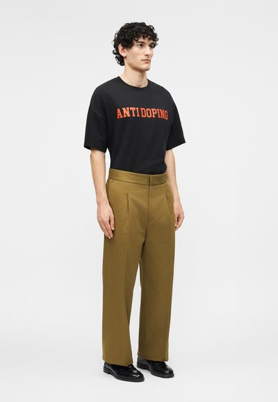 Jeune homme aux cheveux bouclés portant un t-shirt noir "ANTIDOPING", un pantalon marron taille haute à jambes larges et des chaussures habillées noires, debout devant un fond blanc.