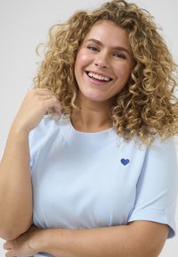 Jasnoniebieska koszulka z krótkimi rękawami, okrągłym dekoltem i małym granatowym serduszkiem haftowanym po lewej stronie. Miękka faktura materiału.