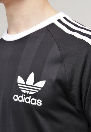 Close-up af en sort Adidas-trøje med hvide striber på skulderen og Adidas-logoet på brystet.