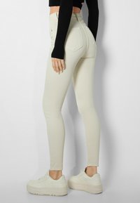 Mujer vestida con vaqueros ajustados de talle alto en color beige y zapatillas de plataforma blancas, de pie de lado contra un fondo de pared lisa.