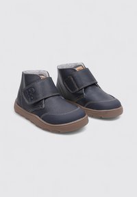 Botas de tobillo para niños negras con suelas marrones y anchas correas de gancho y bucle, con costuras beige visibles sobre un fondo liso.