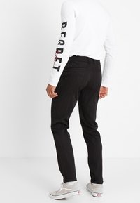 Persoon draagt zwarte gestreepte broek, wit overhemd met lange mouwen met de tekst "REGRET" op de mouw, en beige sneakers, staand tegen een witte achtergrond.